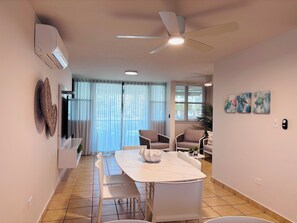 A modern living room with a dining area, a ceiling fan, and a large window with curtains. - Famous beach resort, 5 pools & children's water park / full air conditioning / ocean view (Loiza)