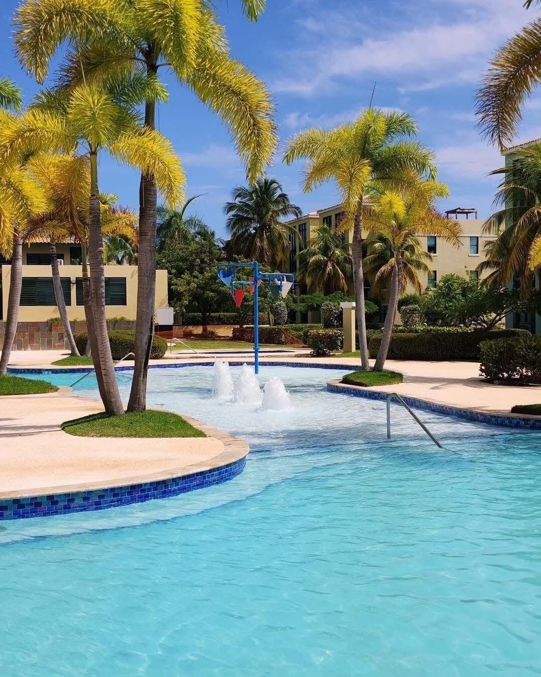 Una piscina con fuente, rodeada de palmeras y un edificio al fondo.
