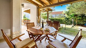 A covered patio with a wooden table and chairs, a bottle of wine, and a bowl of fruit. - Lovely apartment in Banjol with swimming pool (Banjol)