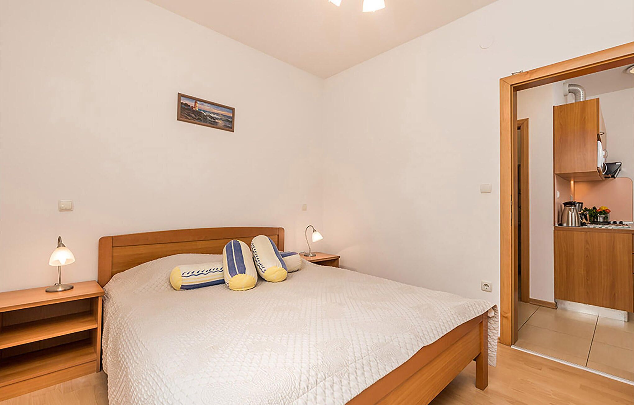 Camera da letto con un letto in legno, due comodini con lampade, un piccolo quadro appeso al muro e vista su un angolo cottura.