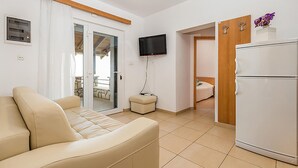 Quarto moderno e bem iluminado com sofá bege, televisão de tela plana na parede, geladeira branca e armário de madeira com um vaso de flores.
