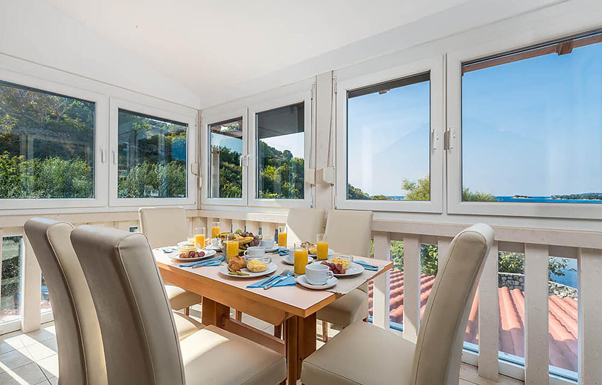 Un coin repas avec une table mise pour le petit-déjeuner, de grandes fenêtres offrant une vue sur la verdure et un plan d’eau, et un balcon doté d’un toit recouvert de tuiles rouges.