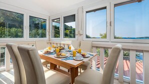 Ein Essbereich mit Tisch, gedeckt für das Frühstück, große Fenster mit Blick auf Grünflächen und ein Gewässer, sowie ein Balkon mit roten Ziegelpfannen.