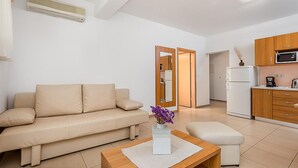 Een moderne woonkamer met een beige bank, een houten salontafel en een kleine witte tafel met een vaas paarse bloemen. In de achtergrond is een keuken met houten kasten en een koelkast zichtbaar.