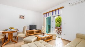 Ein Wohnzimmer mit Sofa, Couchtisch, Fernseher und Esstisch mit Stühlen. Eine Glastür führt zu einem Außenbereich mit Tisch und Stühlen.