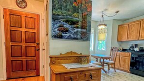 Une table à manger en bois avec des chaises, un coin cuisine avec des meubles en bois et un tableau représentant une scène de forêt accroché au mur.