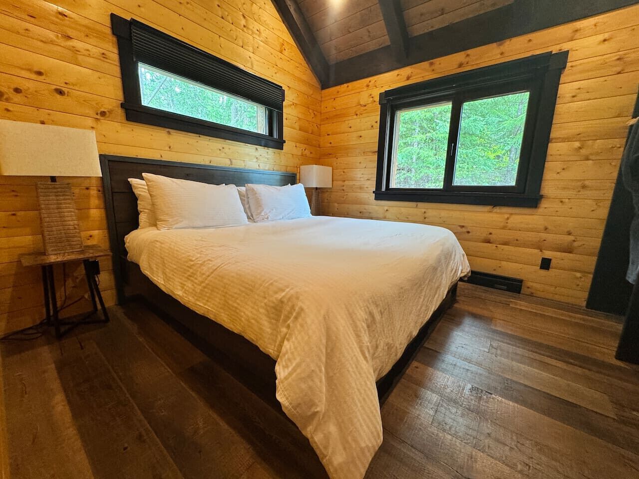 Een slaapkamer in een houten hut, met een bed, twee ramen en een nachtkastje met een lamp.