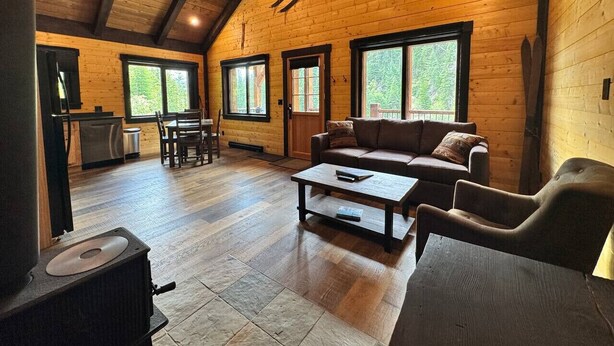 Un acogedor interior de cabaña con detalles de madera, una sala de estar con sofá y mesa de centro, un comedor con mesa y sillas, y una cocina con estufa.