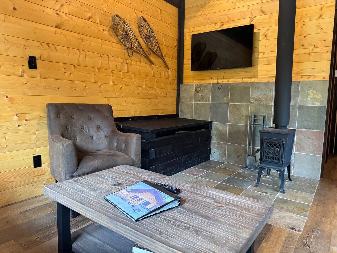 A cozy room with a fireplace, a comfortable armchair, a table with a book, and a television mounted on the wall.
