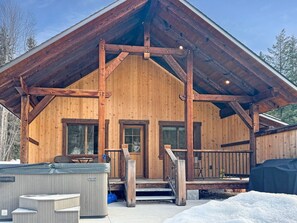 Eine Holzhütte mit überdachter Veranda, einem Whirlpool und verschneitem Gelände.
