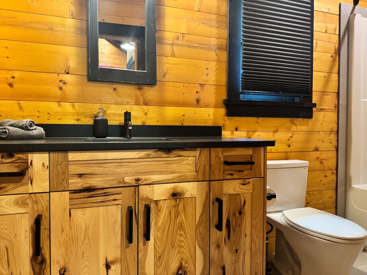 A wooden cabin-style bathroom with a sink, toilet, and window with blinds.