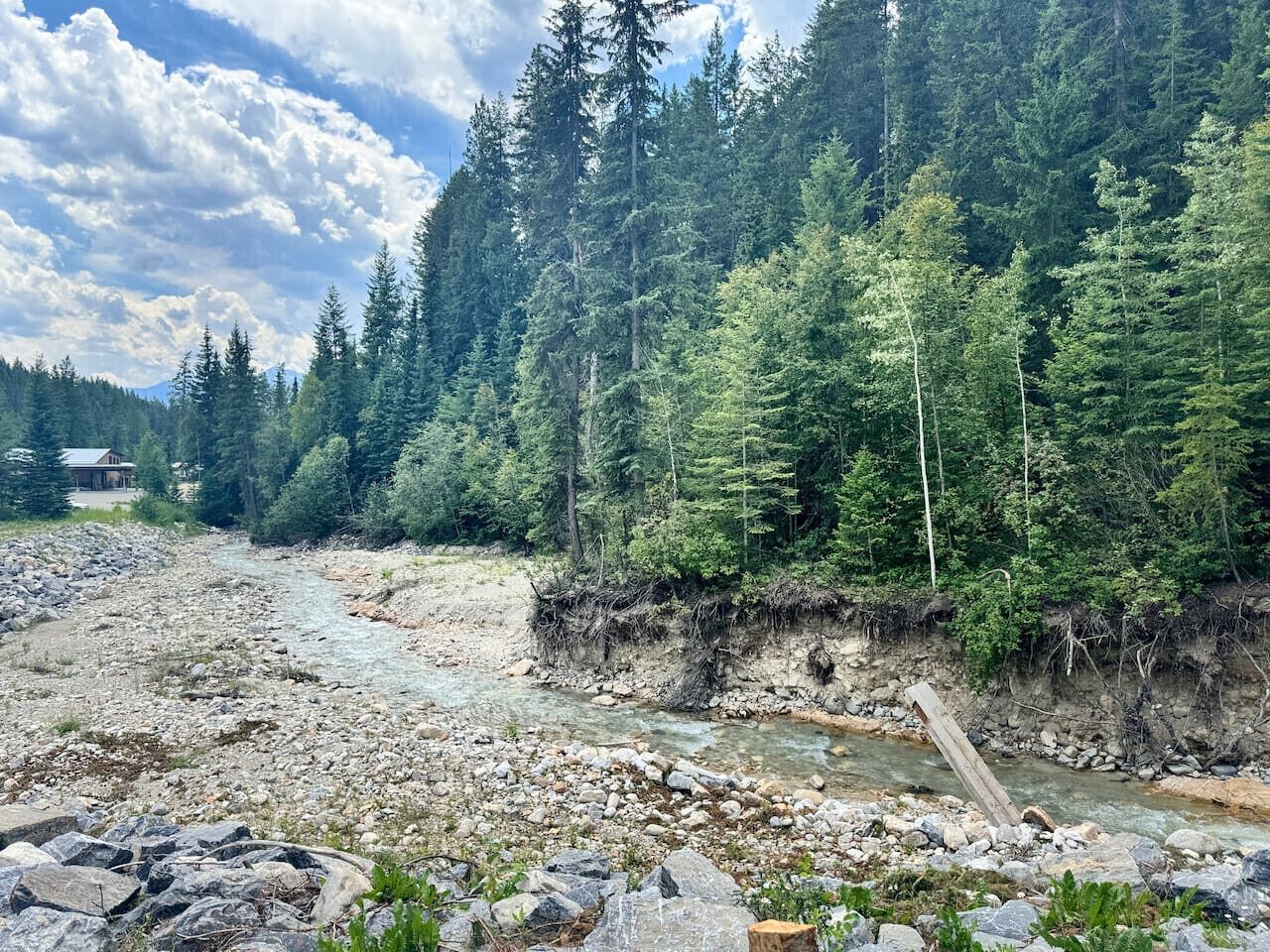 A rocky riverbed with a wooden structure, dense forest, and a house in the background.