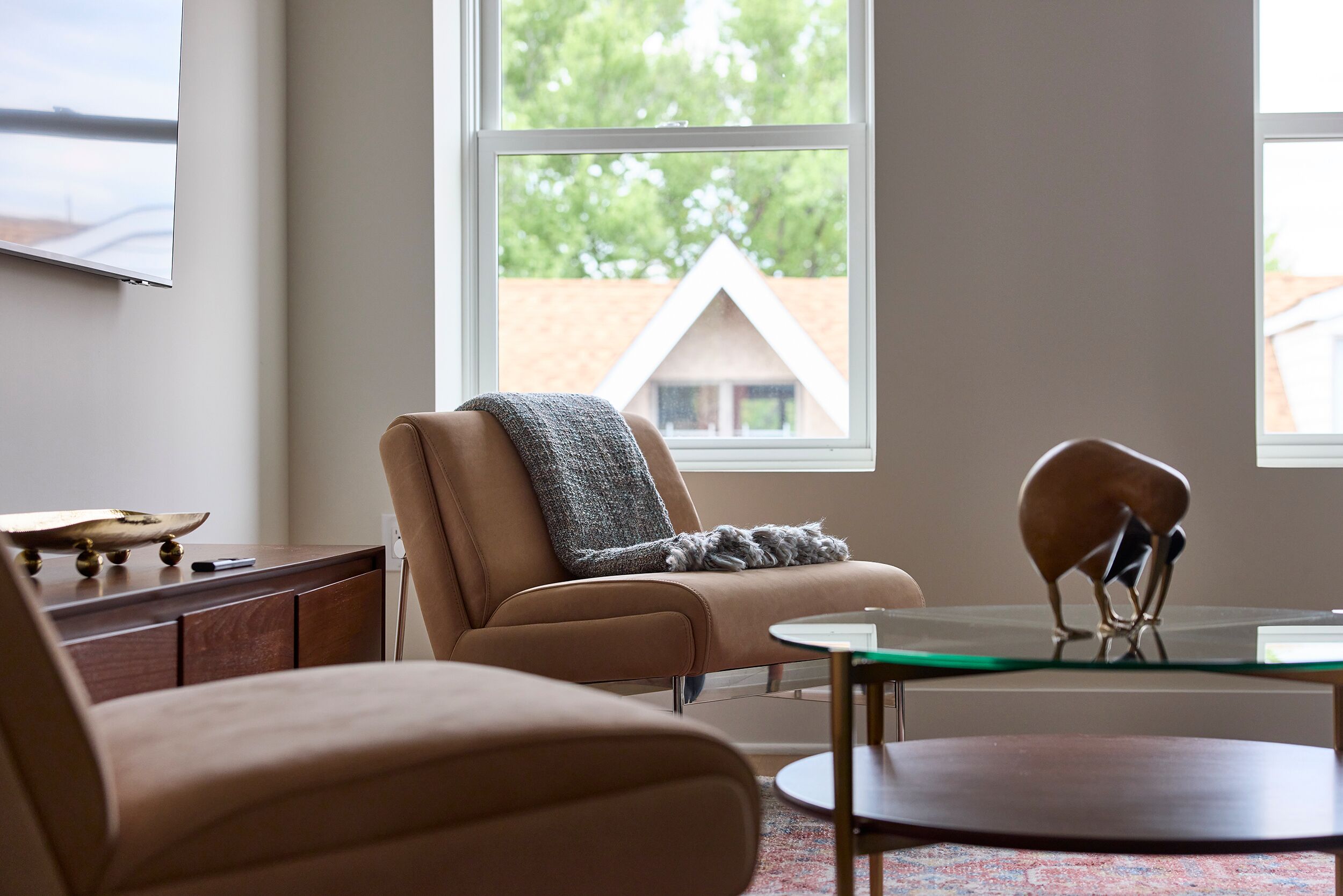 Un salon moderne avec un canapé marron, une table basse en verre et une vue sur une maison par une grande fenêtre.
