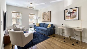 A modern living room with a blue sofa, a white armchair, a glass coffee table, a desk with a chair, and a large abstract painting on the wall. - Sunny 2BR Loft Work Relax Minutes to NYC (Hoboken)