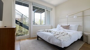 A bedroom with a bed, a wooden nightstand, a window with a view of a staircase, and a ceiling fan. - Hoboken Loft Hideaway - Walk to Path (Hoboken)