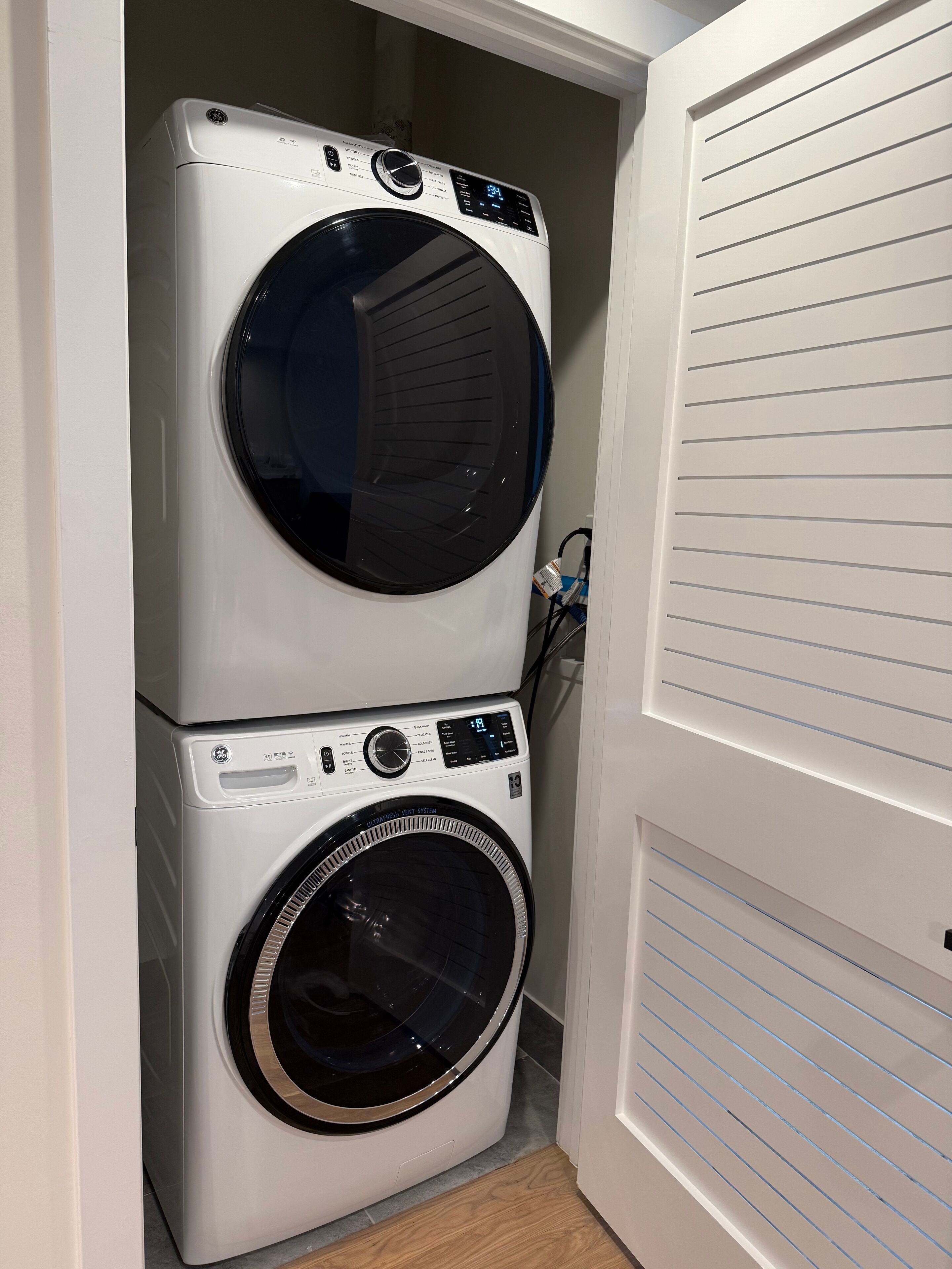 Uma máquina de lavar e secar empilhadas numa lavanderia com uma porta branca e chão ladrilhado.