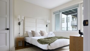 Ein modernes Schlafzimmer mit einem großen Bett, zwei Nachttischlampen, einem Holznachttisch, einer grünen Tagesdecke und Blick auf das gegenüberliegende Gebäude durch ein Fenster.