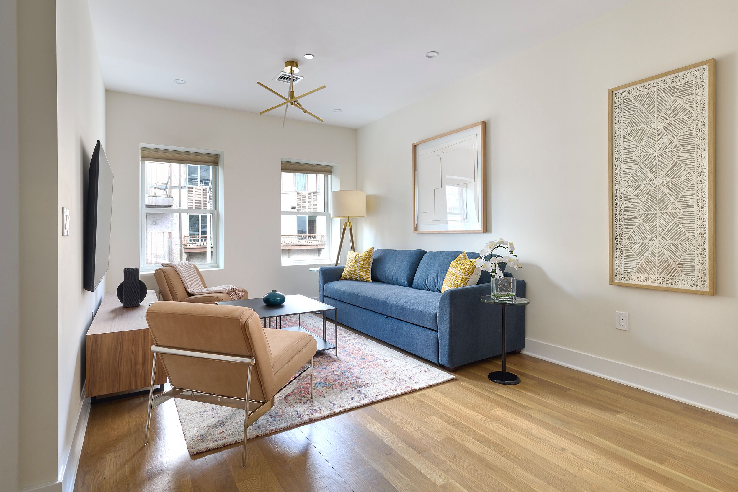 A modern living room with a blue sofa, a beige armchair, a coffee table, and a floor lamp. There are two windows with blinds, a ceiling fan, and a large abstract painting on the wall.