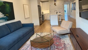 Una sala moderna con un sofá azul, una mesa de centro de vidrio y un sillón beige.