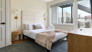 Une chambre moderne avec un grand lit, une table de chevet, une commode, une fenêtre donnant sur la ville et un ventilateur de plafond.