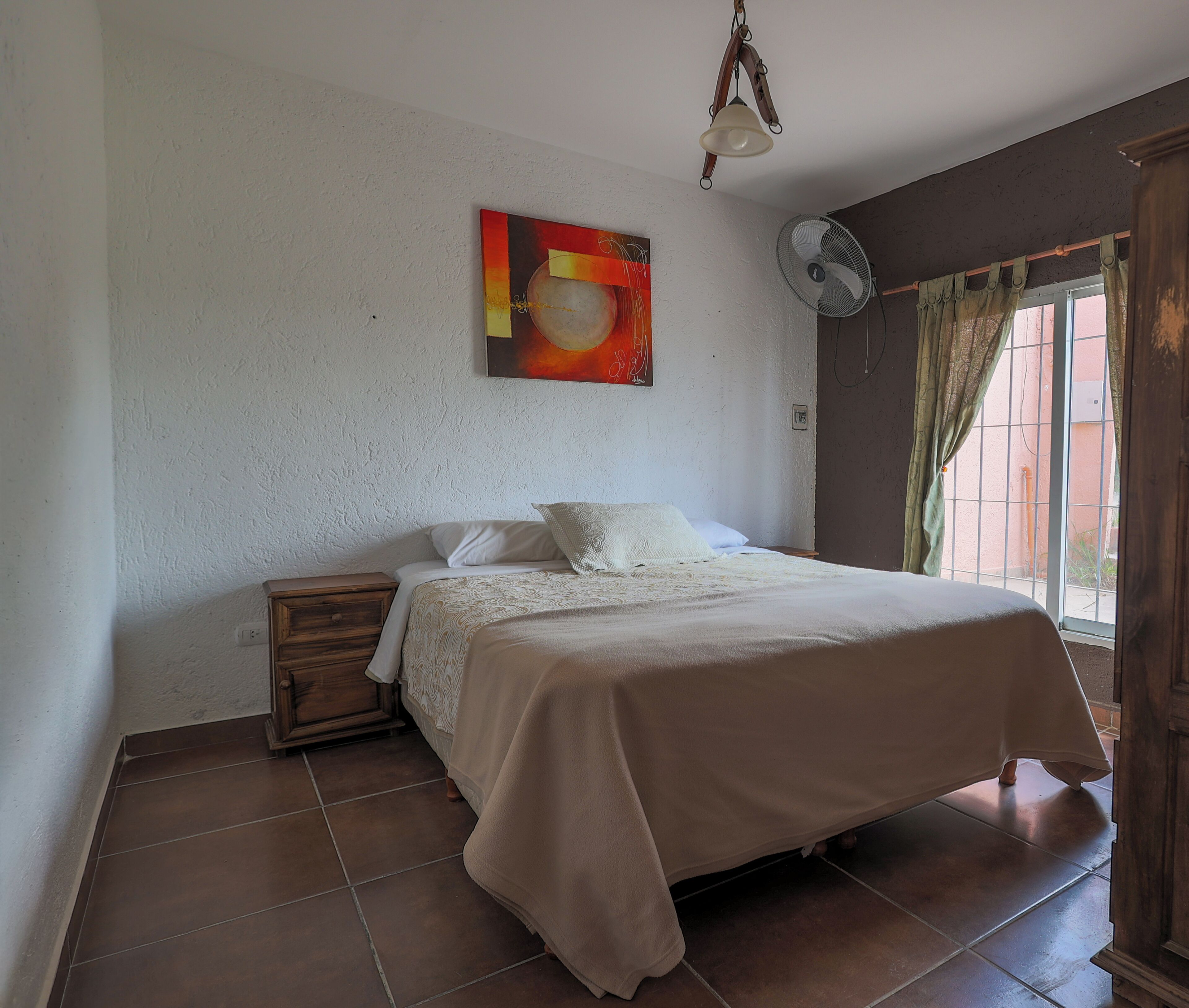 A bedroom with a bed, a nightstand, a wall-mounted fan, a ceiling fan, and a window with curtains.