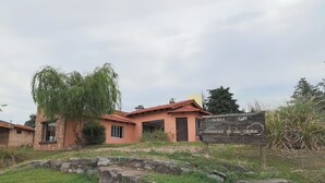 A rustic building with a stone facade and a red-tiled roof, surrounded by greenery and a stone wall.