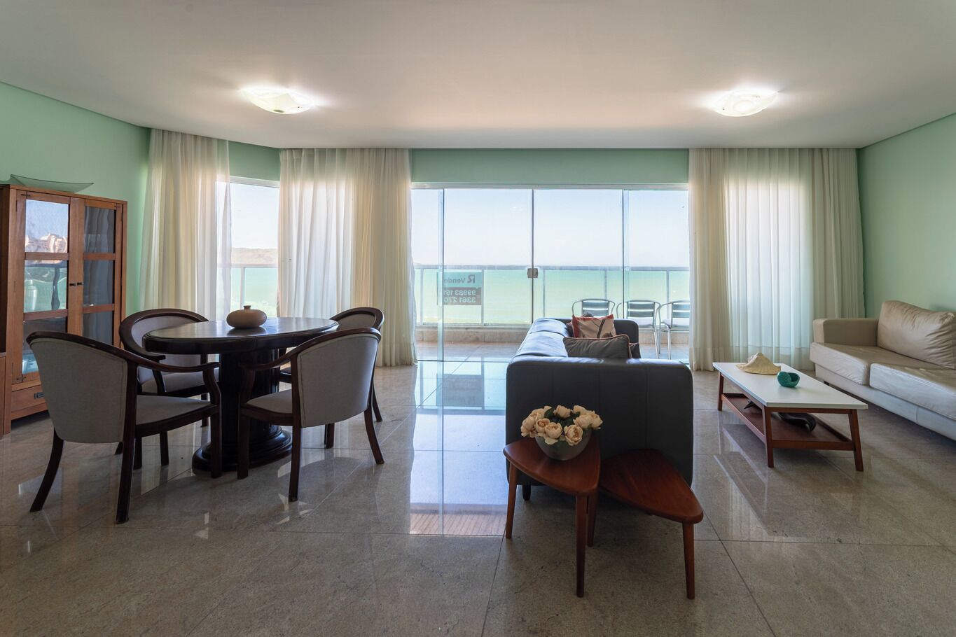 A spacious living area with a dining table, chairs, a sofa, and a coffee table. A large window offers a view of the outdoors.