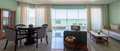 A spacious living area with a dining table, chairs, a sofa, and a coffee table. A large window offers a view of the outdoors.