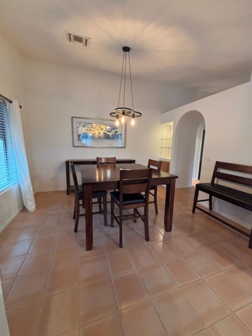 Un espace repas comprenant une grande table, six chaises, un banc, une suspension et une œuvre d’art encadrée accrochée au mur.