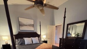 A four-poster bed with a dark headboard, a ceiling fan, a patterned rug, a nightstand with a lamp, and a dresser with a mirror. - The Sleepy Hollow Hideaway (Gold Canyon)