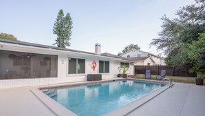 A residential swimming pool with a covered patio area, a wooden fence, and a house in the background.