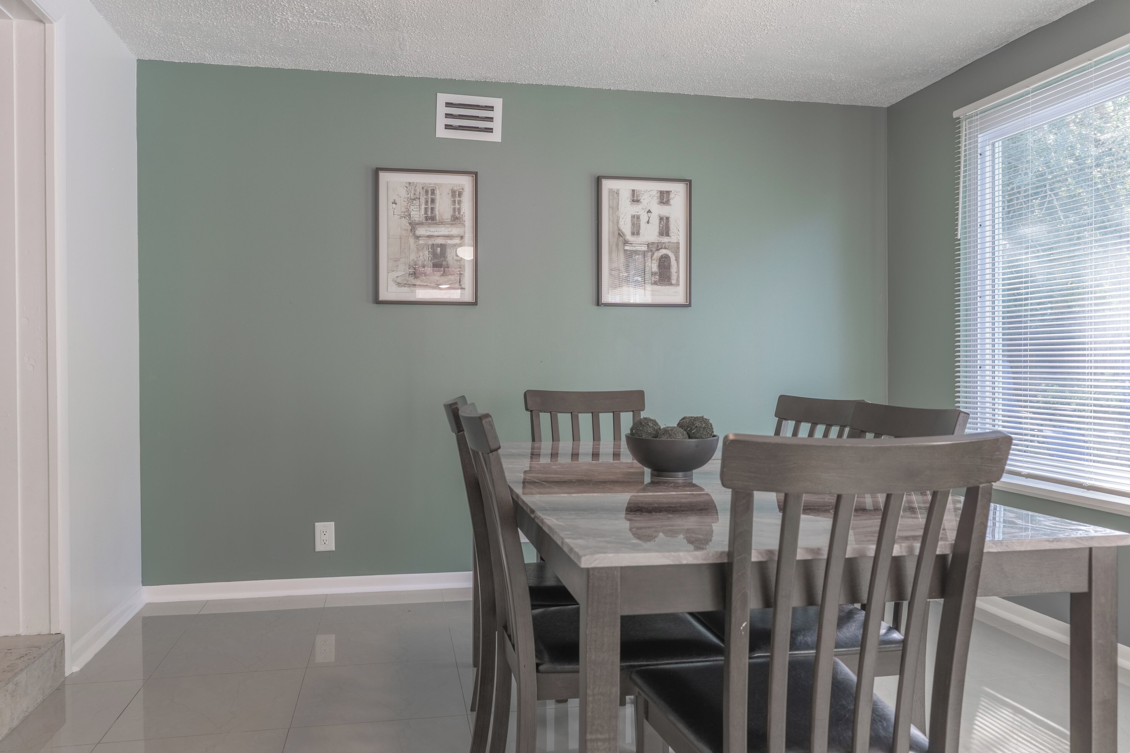 A dining area with a table and chairs, framed pictures on the wall, and a window with blinds.