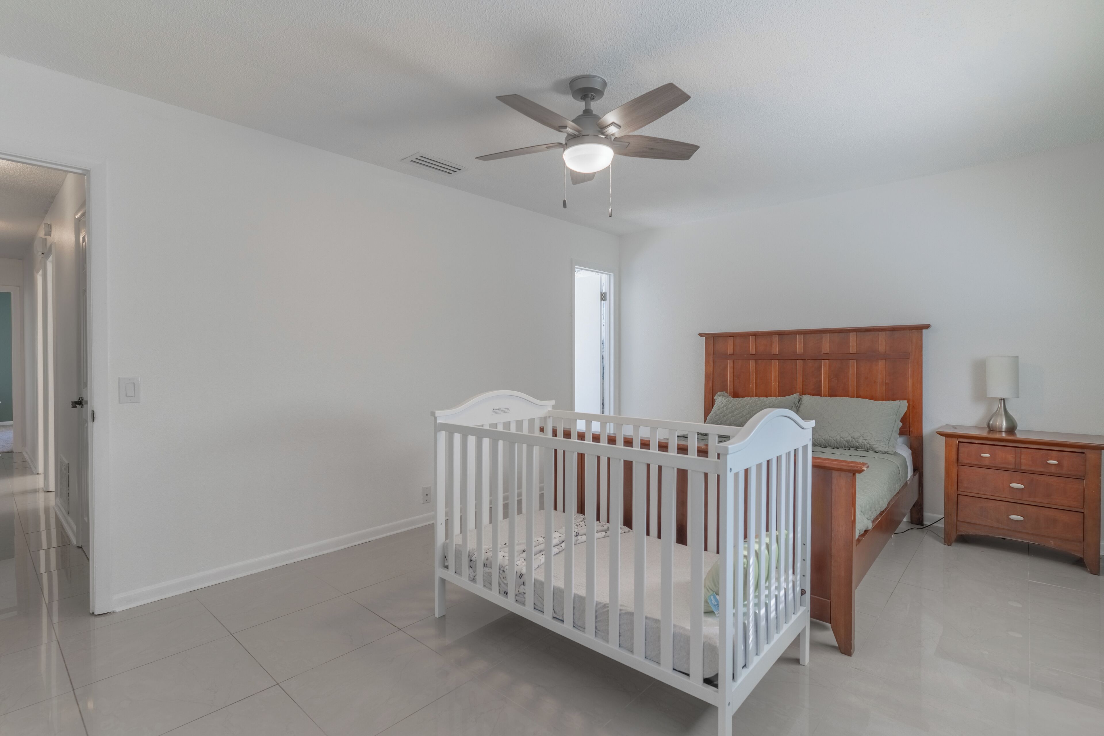 Una habitación amplia y bien iluminada con una cuna blanca, una cama de madera y una mesita de noche.