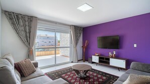 Un salon moderne avec un mur d’accent violet, un téléviseur à écran plat, un canapé, une table basse et une porte-fenêtre coulissante donnant sur un balcon.