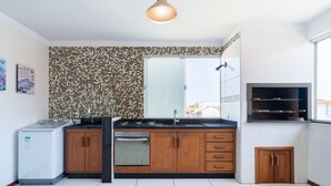 Une cuisine avec un mur recouvert de carreaux, des meubles en bois, un four encastré, un réfrigérateur et une fenêtre avec des stores.