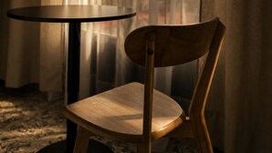 Une chaise en bois et une table ronde dans une pièce donnant sur un paysage que l’on aperçoit à travers de fines rideaux.