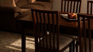 Un salon avec un canapé marron, des chaises en bois, une table à manger ornée de fruits et un téléviseur fixé au mur.