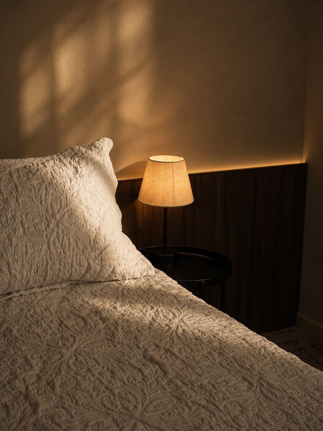 Una cama con edredón blanco texturizado, una lámpara de noche y una mesita de noche de madera oscura.