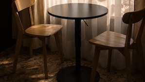 Une pièce avec une table ronde et deux chaises en bois, une fenêtre ornée de rideaux légers, et une vue sur des immeubles et des arbres.