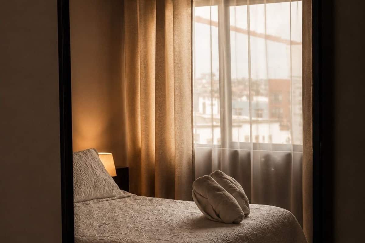 A hotel room with a bed, a ceiling fan, a window with curtains, and a view of buildings outside.