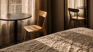 Une chambre d’hôtel avec un lit, deux chaises, une table ronde, un grand miroir et des rideaux légers.