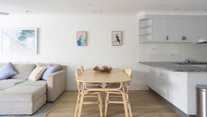 Een moderne woonkamer met een beige bank, een houten eettafel met vier stoelen, en een keuken met witte kasten en een grijs aanrecht.
