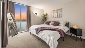 Une chambre à coucher avec un grand lit, une table de chevet, une lampe sur pied, une fenêtre avec des rideaux et un tableau au mur.