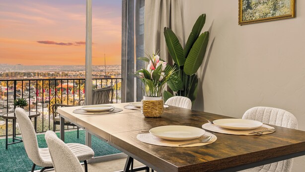 Une table à manger dressée avec des assiettes, un vase rempli de fleurs et des chaises, le tout dans une pièce offrant une vue sur le paysage urbain.