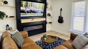 A modern living room with a large flat-screen TV mounted on a black wall, a ceiling fan, and a fireplace. There are two brown leather sofas and a coffee table with a plant. - Redbud: Waterfront, Augusta Canal, 3BR, king bed (Augusta)