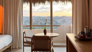 Ein Zimmer mit einem Bett, einem Esstisch mit Stühlen, einem kleinen Tisch mit einer Vase und Blick auf eine Stadtlandschaft durch große Fenster.
