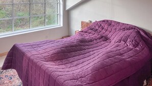 A bedroom with a purple bedspread, a window with a view of trees, and a small bedside table with a lamp.
