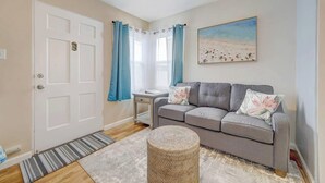 Ein Wohnzimmer mit einem grauen Sofa, einem kleinen Beistelltisch, einem Weidenhocker, einem Streifenmuster-Teppich und einer Tür mit Griff.