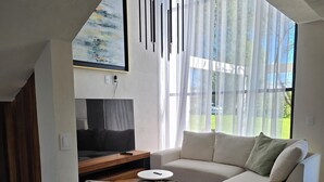 Una sala de estar moderna con un sofá seccional, un sillón verde y una pequeña mesa redonda. Hay un televisor de pantalla plana sobre un soporte de madera, y se aprecia un cuadro en la pared.