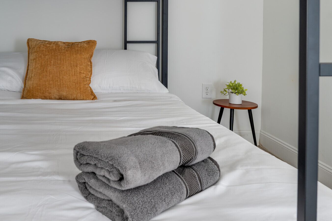 Ein ordentlich gemachtes Bett mit weißen Leinen und einem gefalteten grauen Handtuch, einem senfgelben Kopfkissen und einem kleinen runden Tisch mit einer Pflanze daneben.
